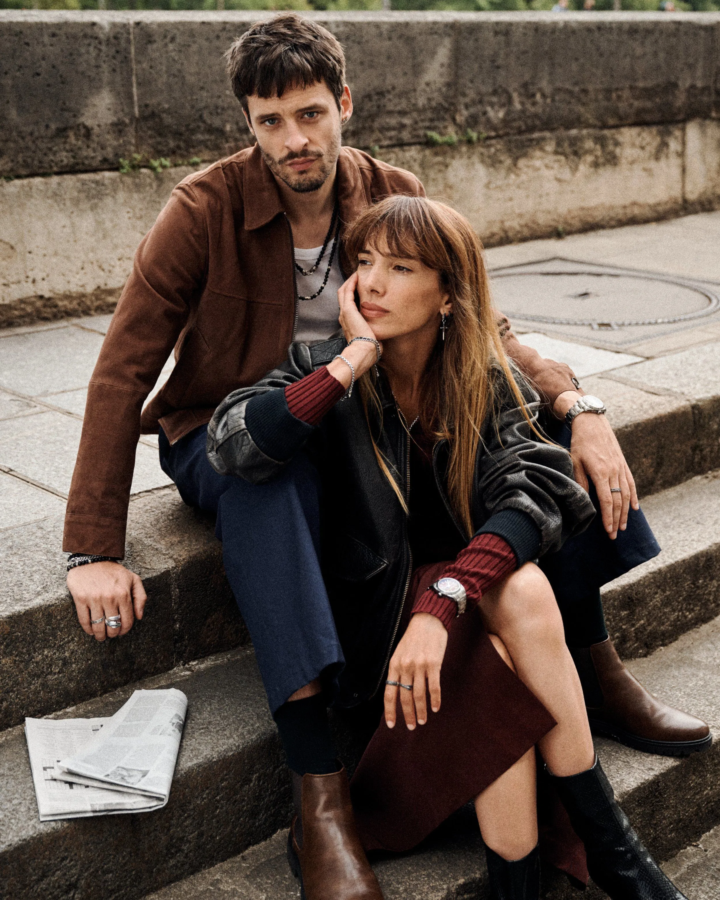 A man and woman sitting on concrete steps outdoors, the man looking directly at the camera and the woman resting her head on his knee, with a newspaper on the steps near them.
